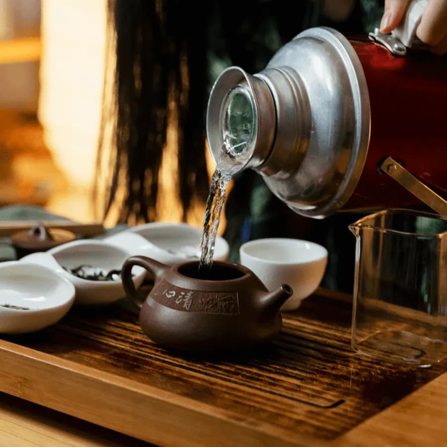 Hot water poured from a thermos into a small clay teapot, beside white cups and dishes of tea leaves