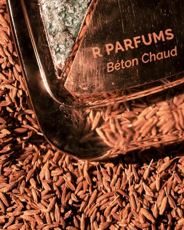 刻有 R Parfums 字样的 Béton Chaud 瓶身一角，静躺在干燥的孜然籽上，映着温暖的铜色光线。