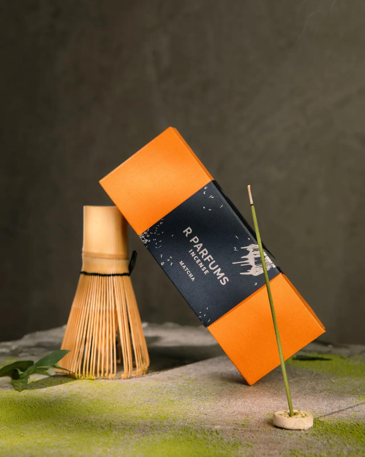 R Parfums Incense Matcha box leaning on a bamboo tea whisk, matcha powder dusted on stone