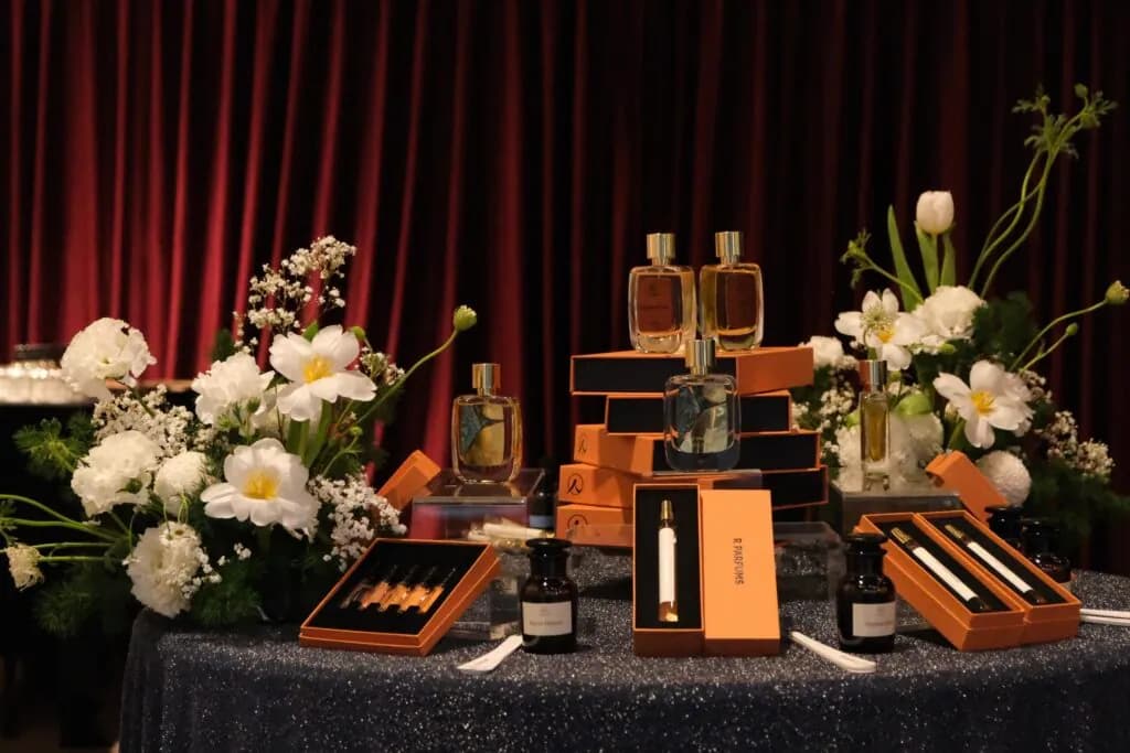 展示桌上，R Parfums 香水瓶与橘色纸盒摆在白花旁，背景是红丝绒布幕，摄于“Light and Shadow”展场。