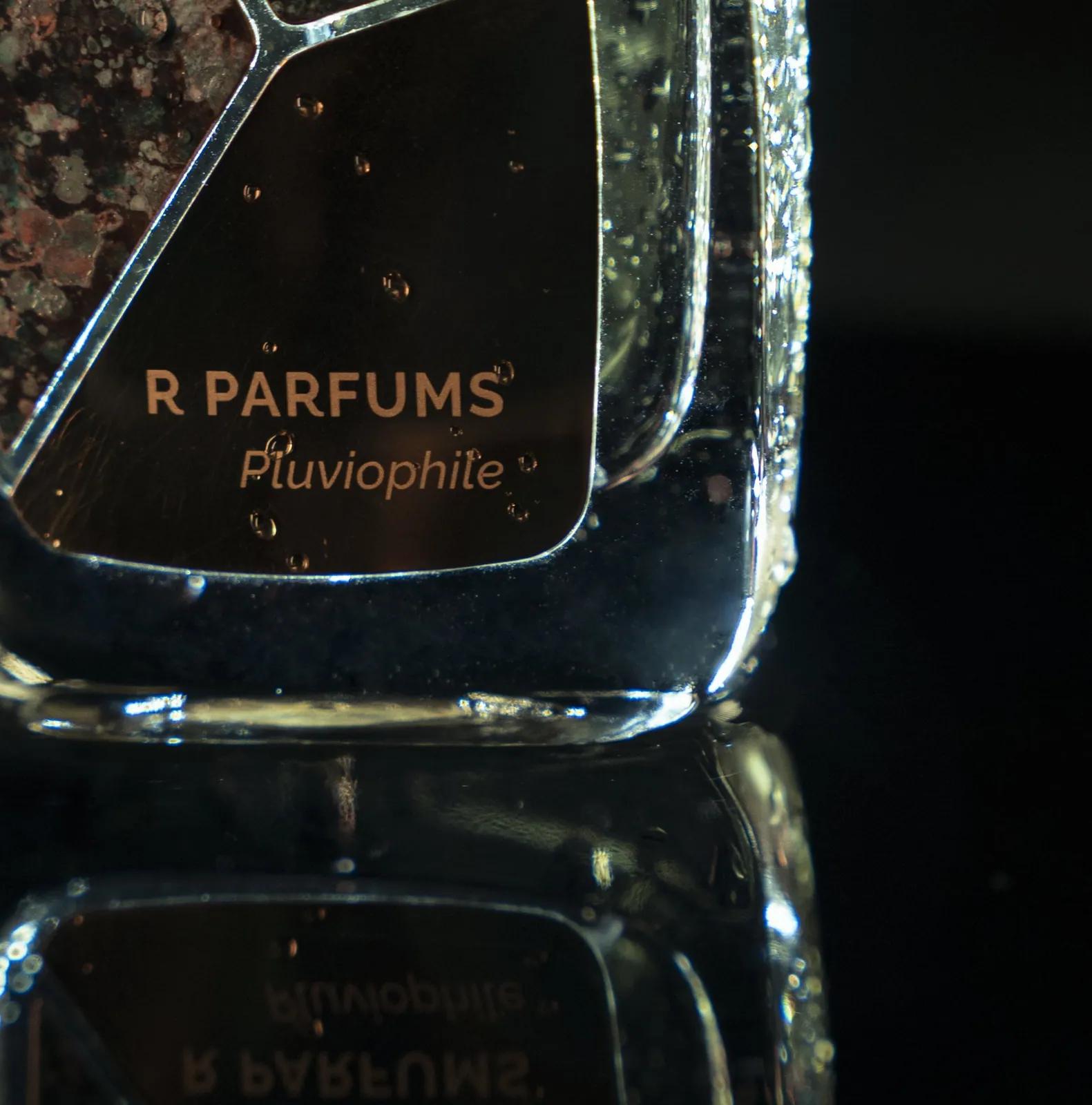 Pluviophile 瓶身一角的特写:黄铜片上的 R Parfums 字样,细小的水珠,瓶影映在深色玻璃上。