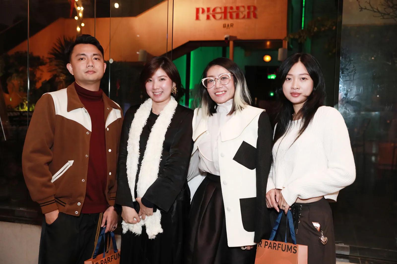 四位来宾在酒吧入口前合影，其中两人提着橘色 R Parfums 纸袋，摄于展览开幕夜。