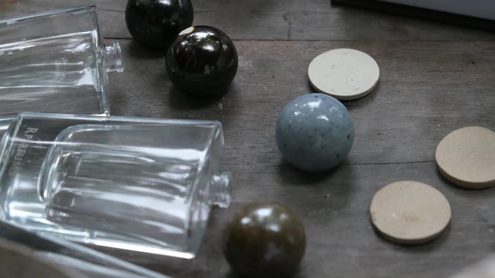A clear glass bottle beside finished ceramic caps in blue-grey, brown and glossy black glaze, with a ceramic charm