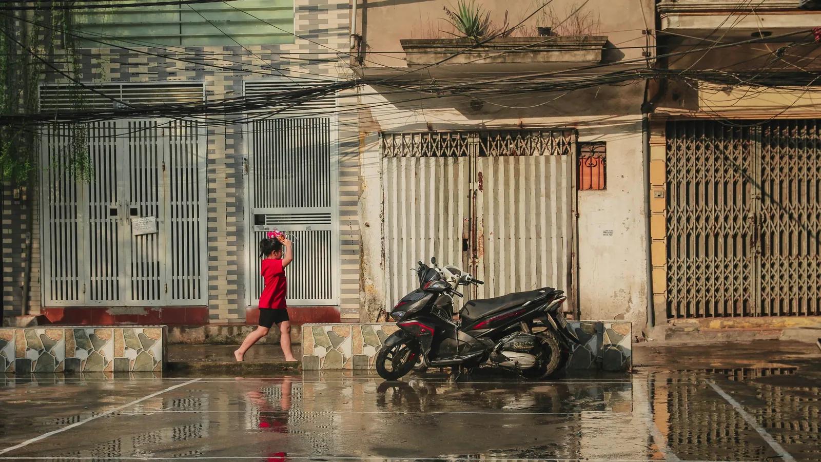 河内街区雨后,穿红衣的小女孩赤脚走在湿漉漉的人行道上,摩托车停在一排铁卷门前。