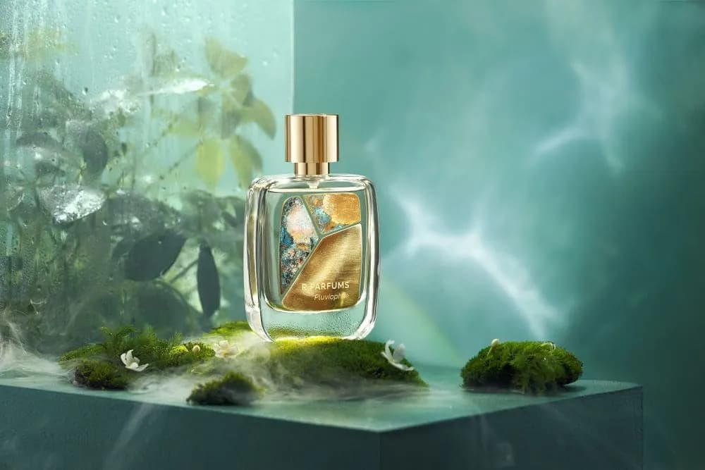 Chai Pluviophile của R Parfums, mặt chai gắn tấm đồng thau patina, đặt trên rêu giữa sương ẩm và lá sau kính mưa.