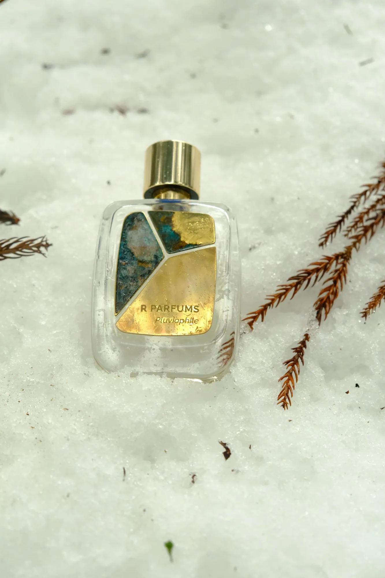 A Pluviophile Patina bottle in snow, close: gold cap, a piece of blue-green patina, a piece half brass and half patina, and a piece of bright brass printed R PARFUMS Pluviophile, sprigs of russet conifer to the right