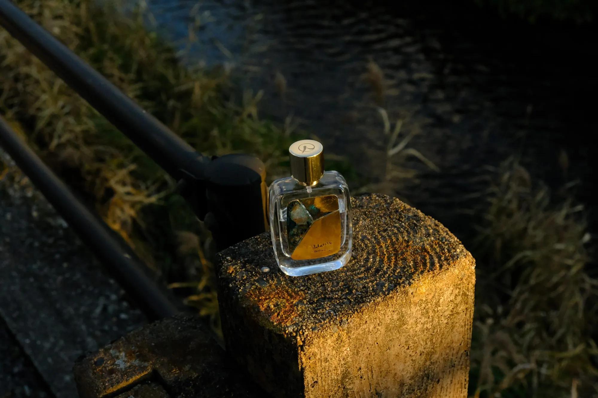 A Patina bottle with a gold cap on top of a rough stone post, lit by low sun; a dark railing to the left, dry grass and dark water behind