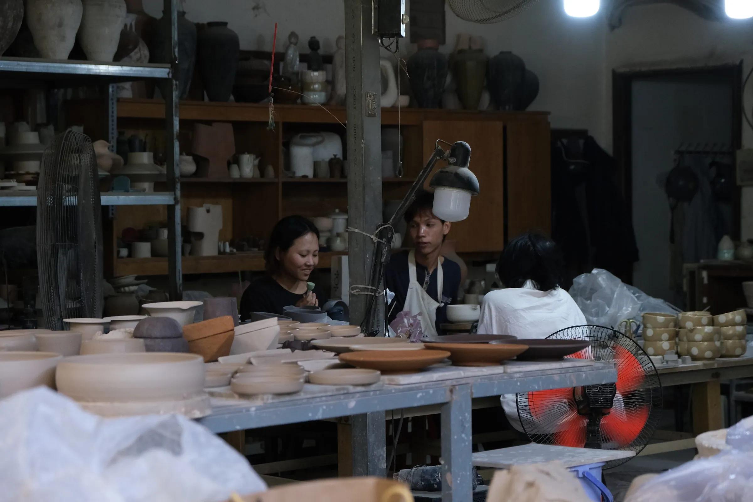 Toàn cảnh xưởng gốm: hai người làm việc bên bàn dài, kệ gốm phía sau