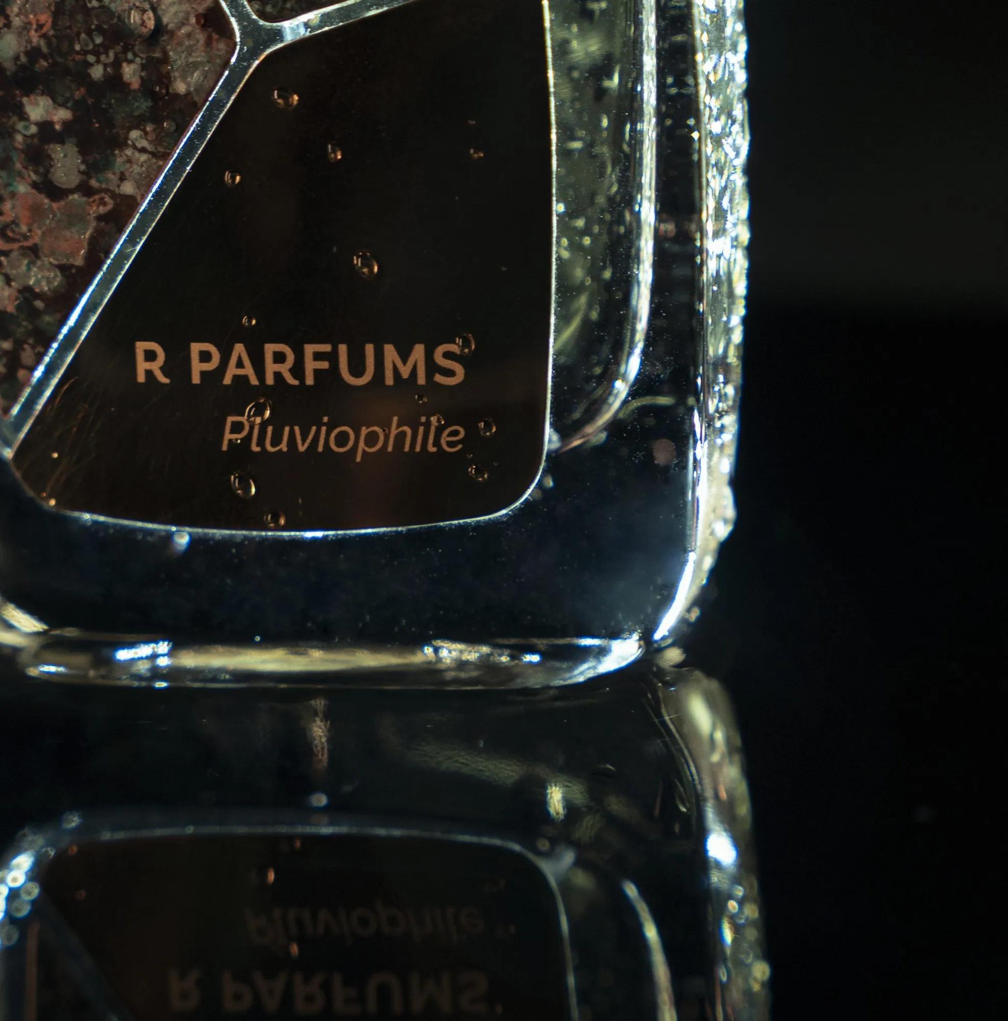 Cận cảnh chai Patina Pluviophile: dòng chữ «R PARFUMS Pluviophile» trên mảnh đồng, giọt nước trên thân chai, bóng chai in trên mặt đen bên dưới