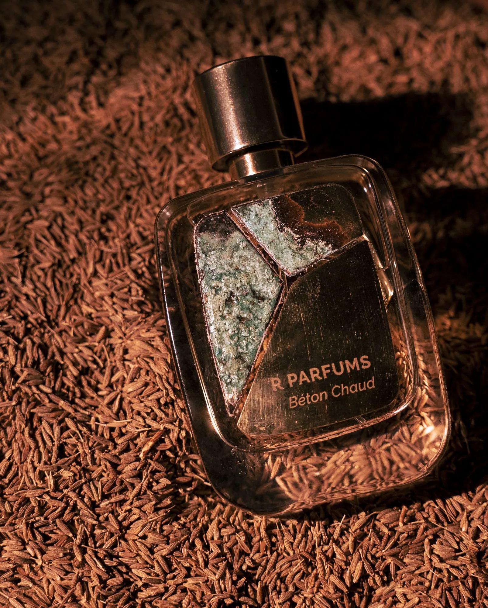 Béton Chaud trong chai Patina nằm trên lớp hạt nhỏ màu nâu, patina xanh bạc trên hai mảnh, dòng chữ «R PARFUMS Béton Chaud» trên mảnh thứ ba