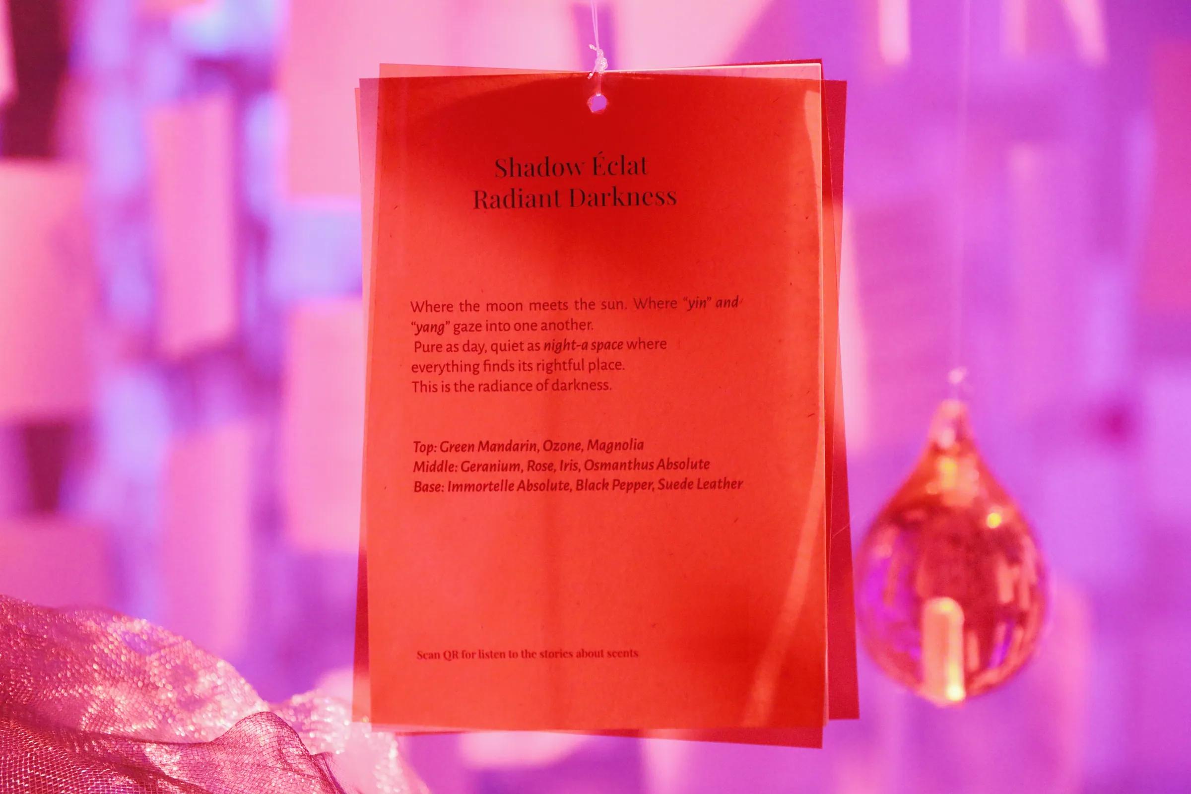 The red Shadow Éclat – Radiant Darkness card hanging beside an orange glass droplet, against pink-violet hanging letters