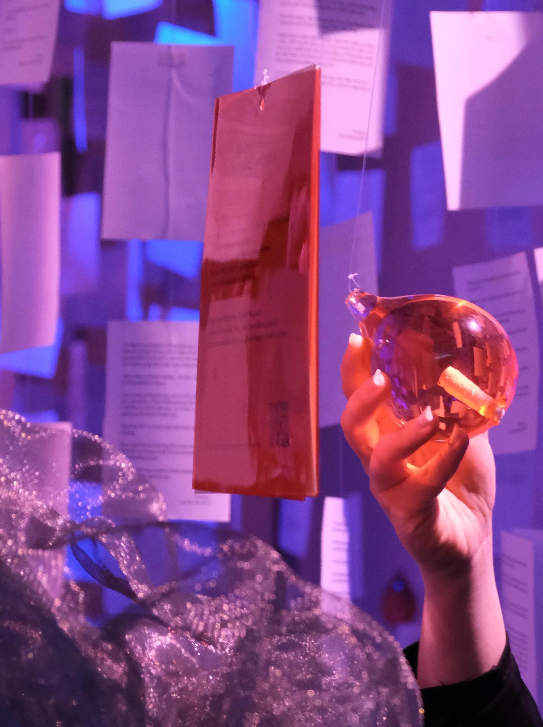 A hand holding up an orange glass droplet beside a red card, among the hanging letters in violet light