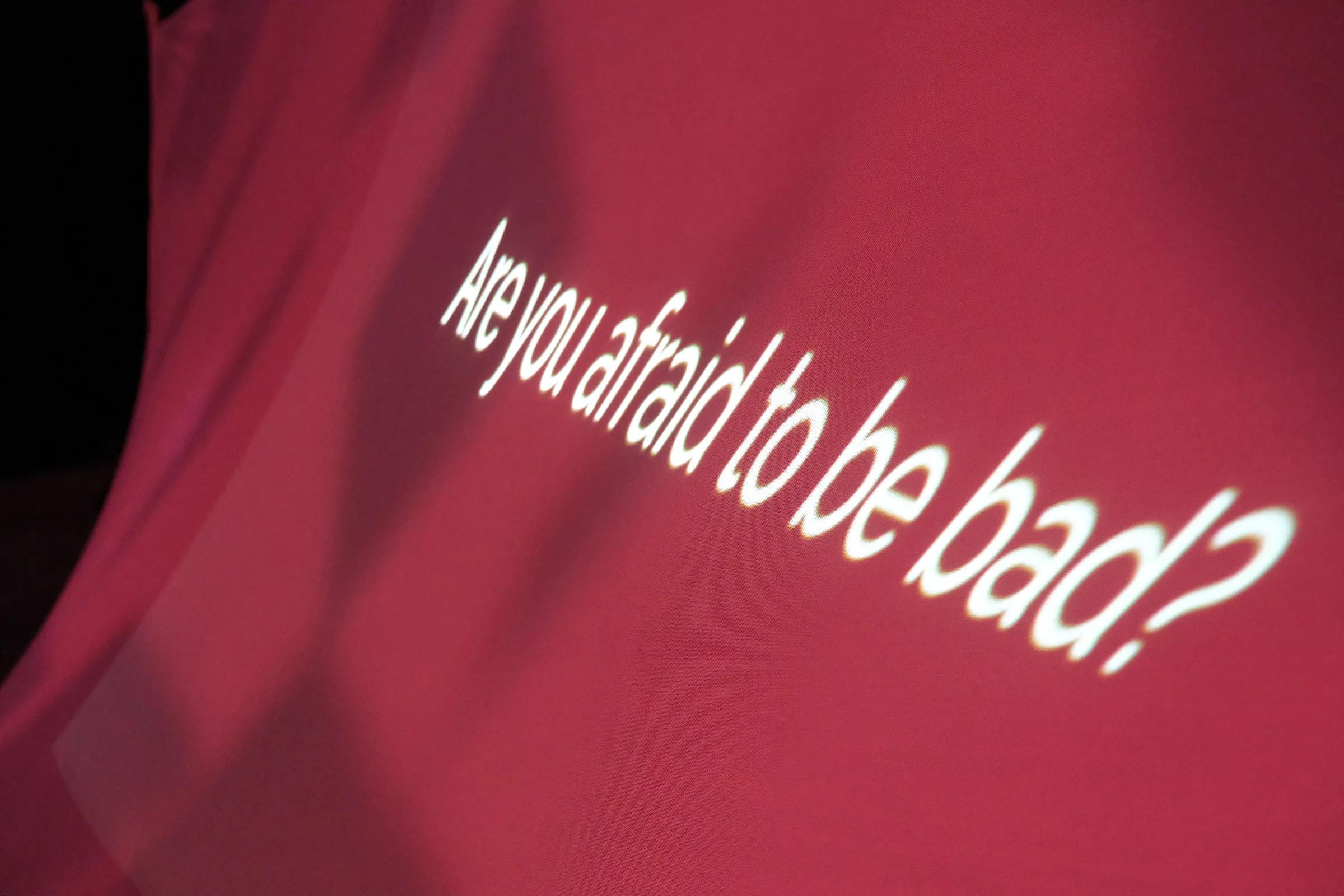 The projected words “Are you afraid to be bad?” on red fabric