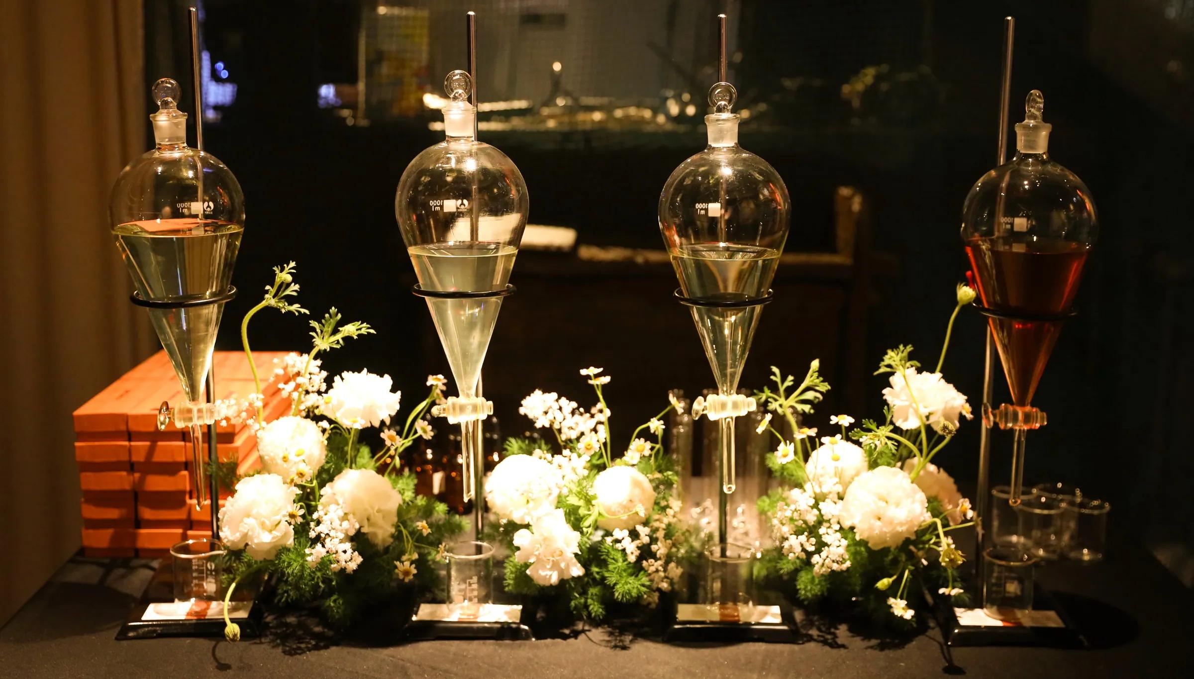 Four glass separating funnels holding scented liquid from pale to amber, standing among white flowers against a dark background, on the private opening night