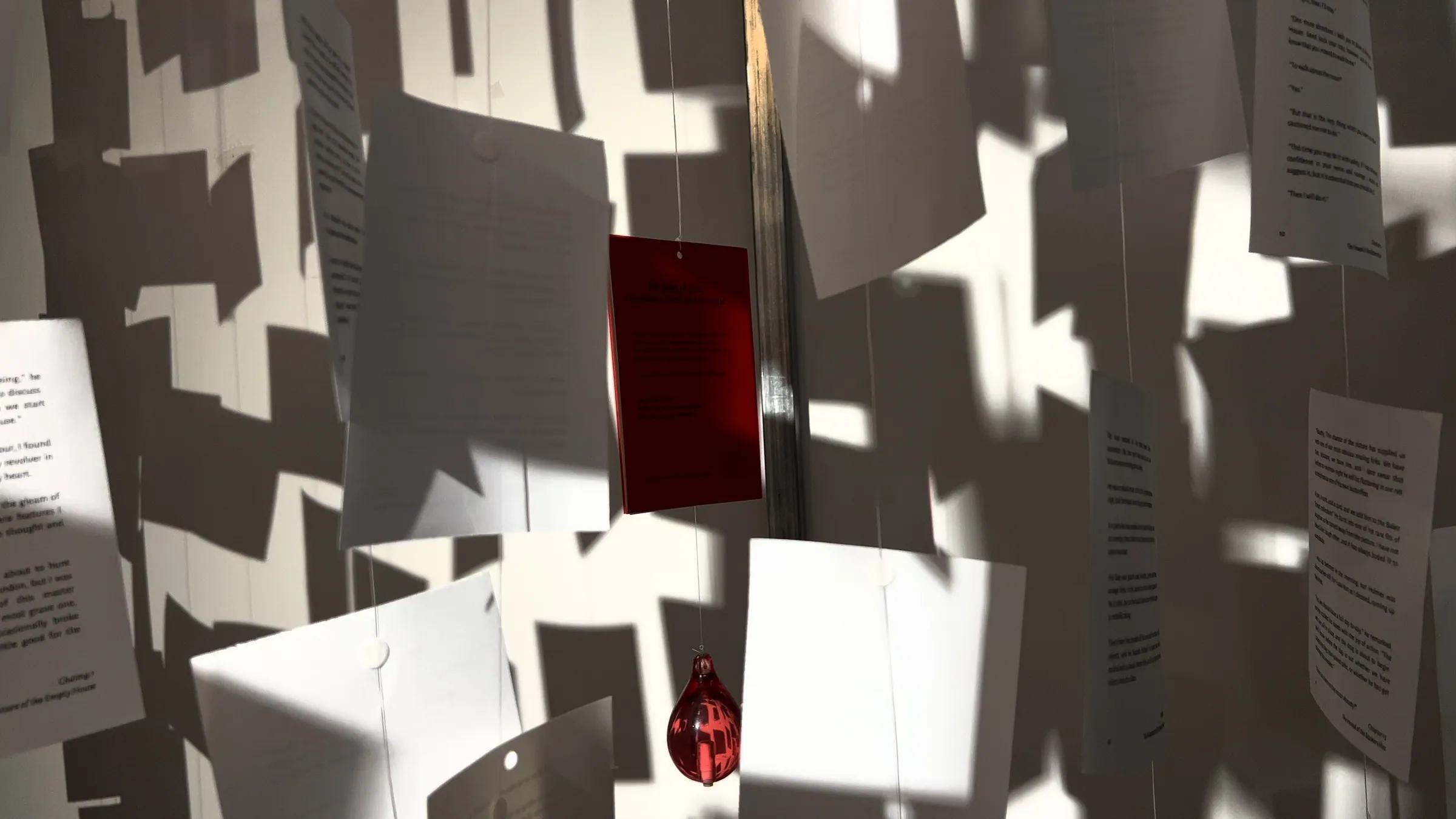 Letter pages, a red card and a red glass droplet hanging in sunlight while the room was being built, their shadows covering the white wall