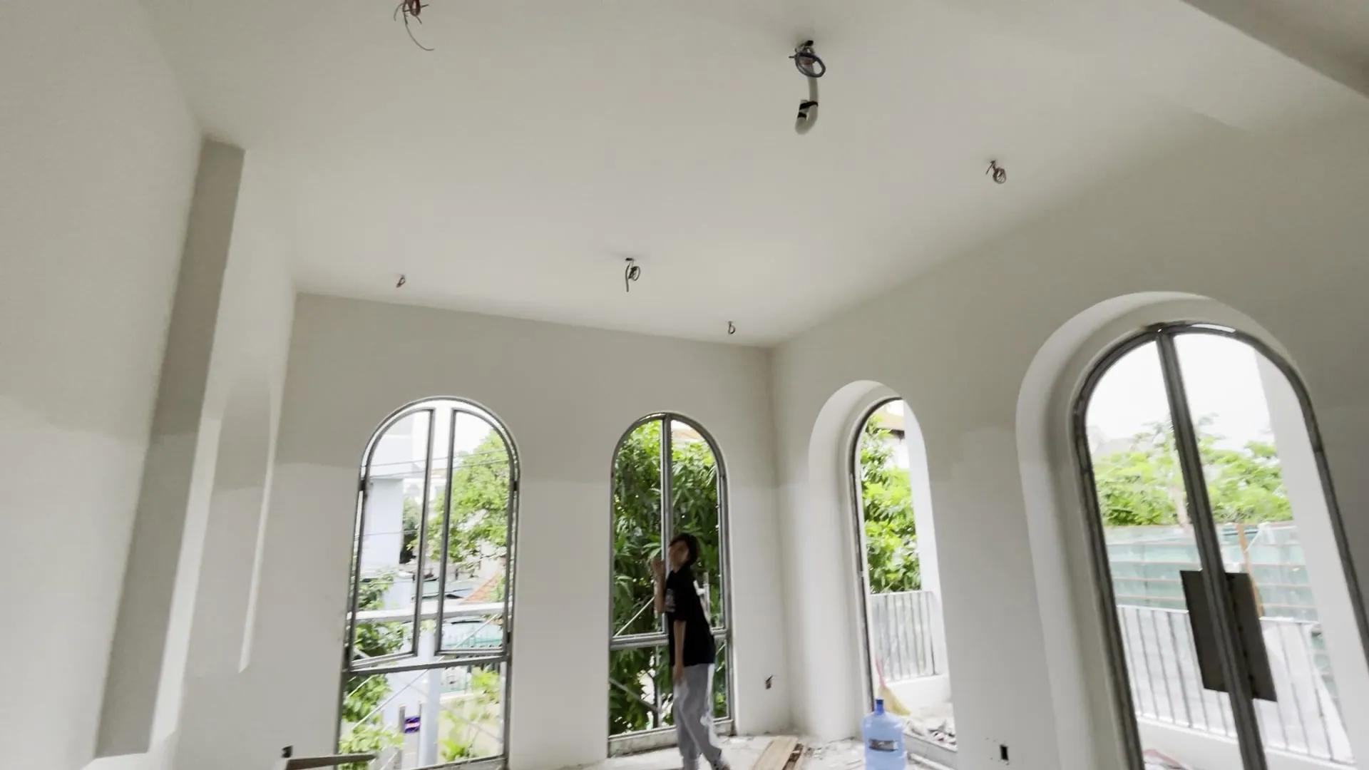 A white room with a sloping ceiling, wires waiting for lights, a row of tall arched windows looking out on greenery; a person stands in the middle of the room