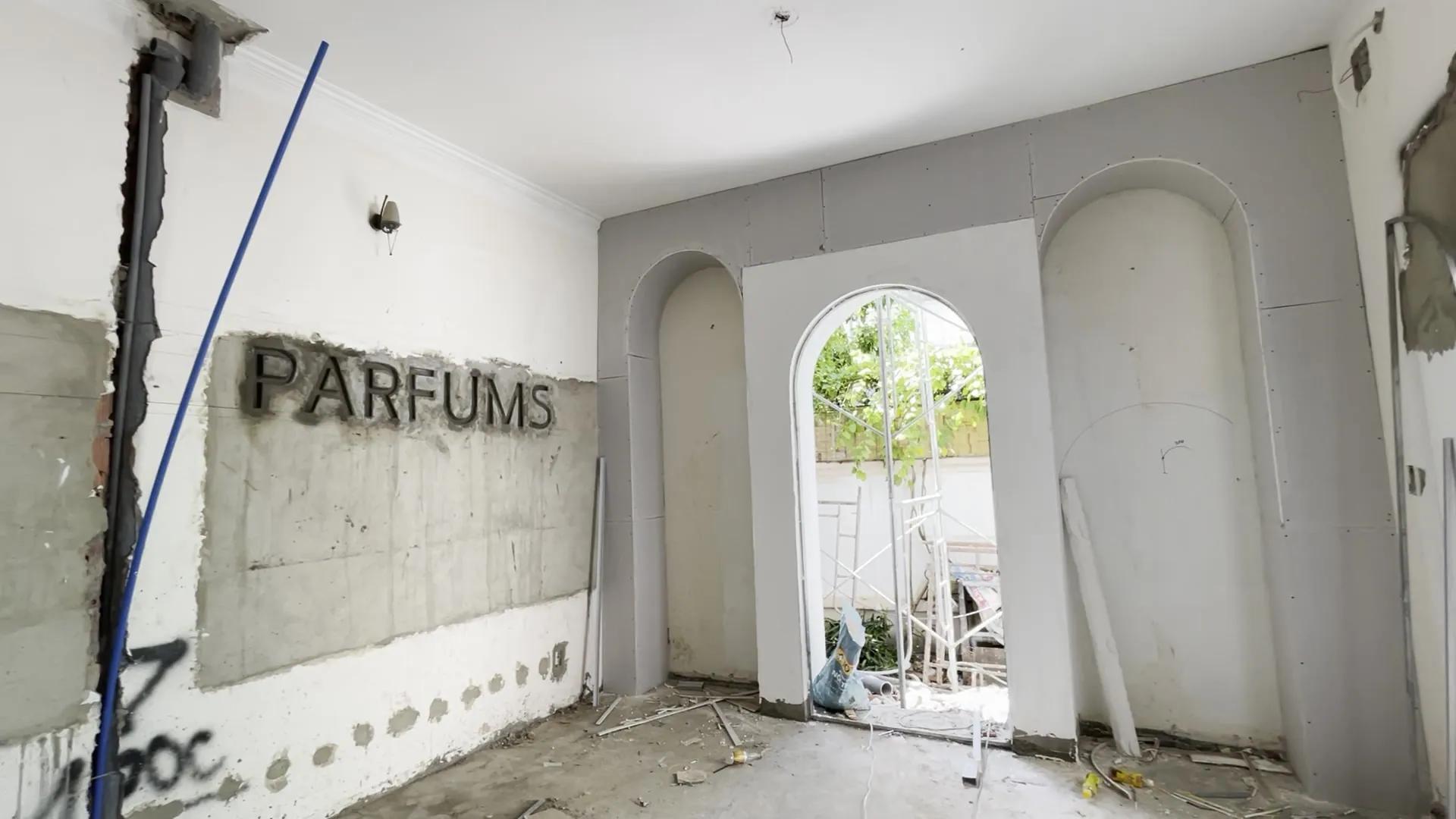 Metal letters spelling PARFUMS on a raw concrete wall; in the middle of the room a white arch opens onto greenery, an arched niche on either side, electrical conduit still running along the wall