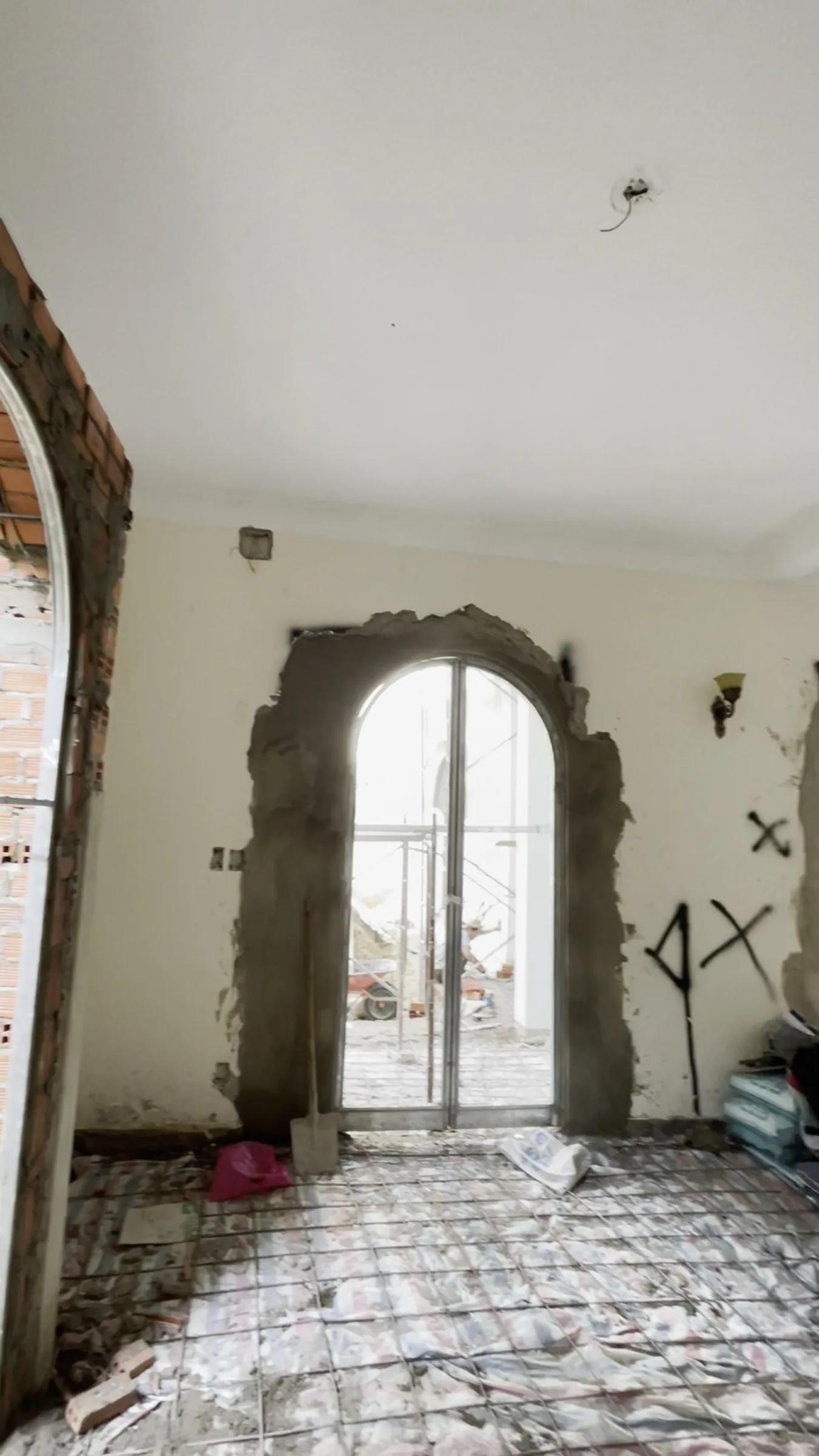 A room under renovation: an arched opening newly widened, its edges raw grey mortar, an arched glass frame standing inside; an X sprayed in paint on the wall, rubble still on the floor