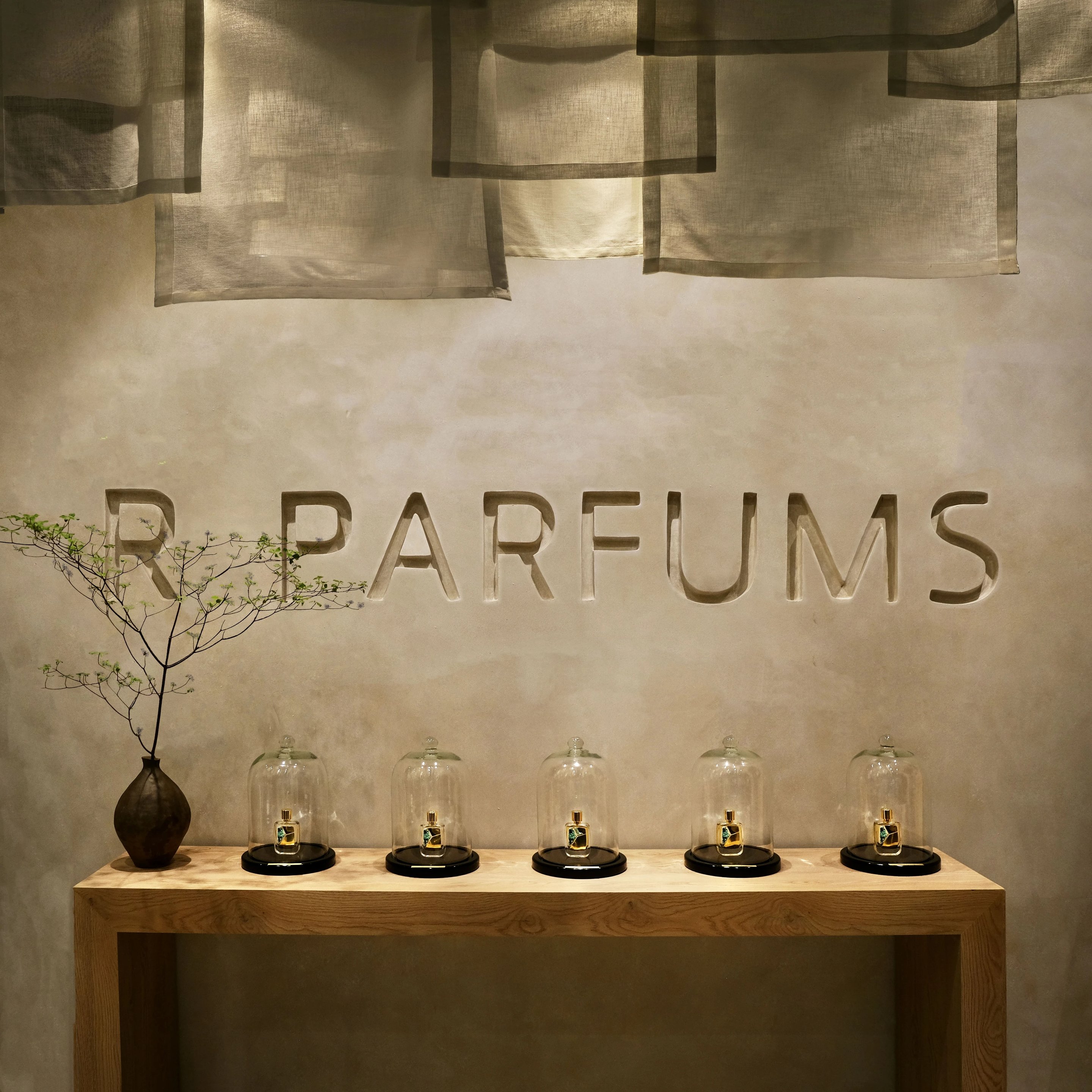R PARFUMS na inukit sa pader na yeso sa itaas ng limang bote sa ilalim ng mga salaming hugis-kampana sa isang mesang kahoy, katabi ng isang plorerang seramika
