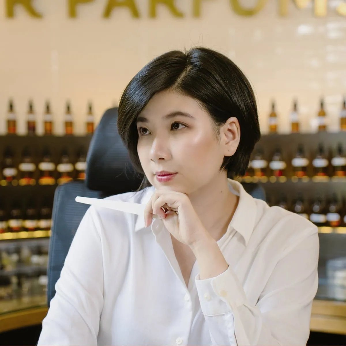 Pencipta wangian Rei Nguyen memegang jalur ujian kertas, dengan rak botol bahan mentah dan tulisan R Parfums pada dinding di belakangnya