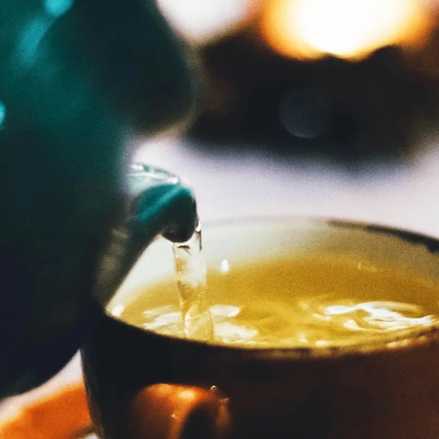 Gambar dekat teh hijau keemasan dituang dari teko ke dalam cawan, berlatarkan bokeh hangat yang lembut