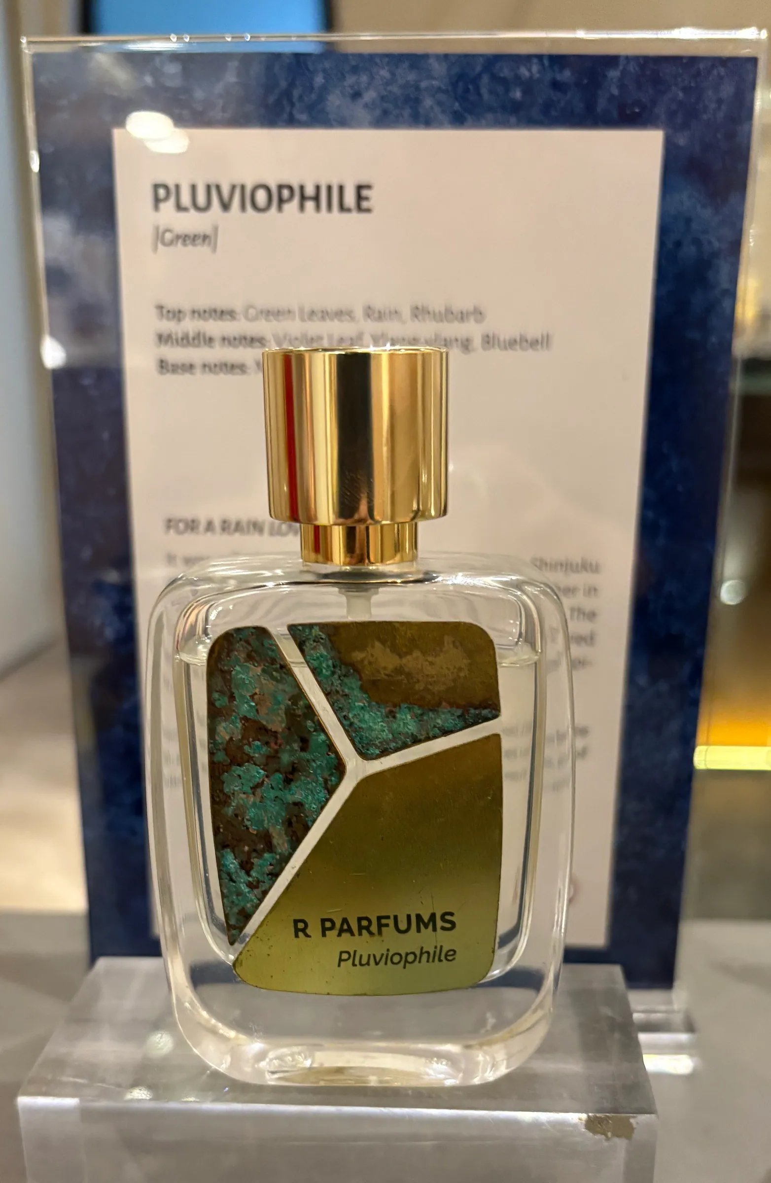 Sebotol Pluviophile Patina di atas blok akrilik jernih, kepingan loyangnya berpatina hijau dan coklat, di hadapan kad yang menyenaraikan nota wangian itu
