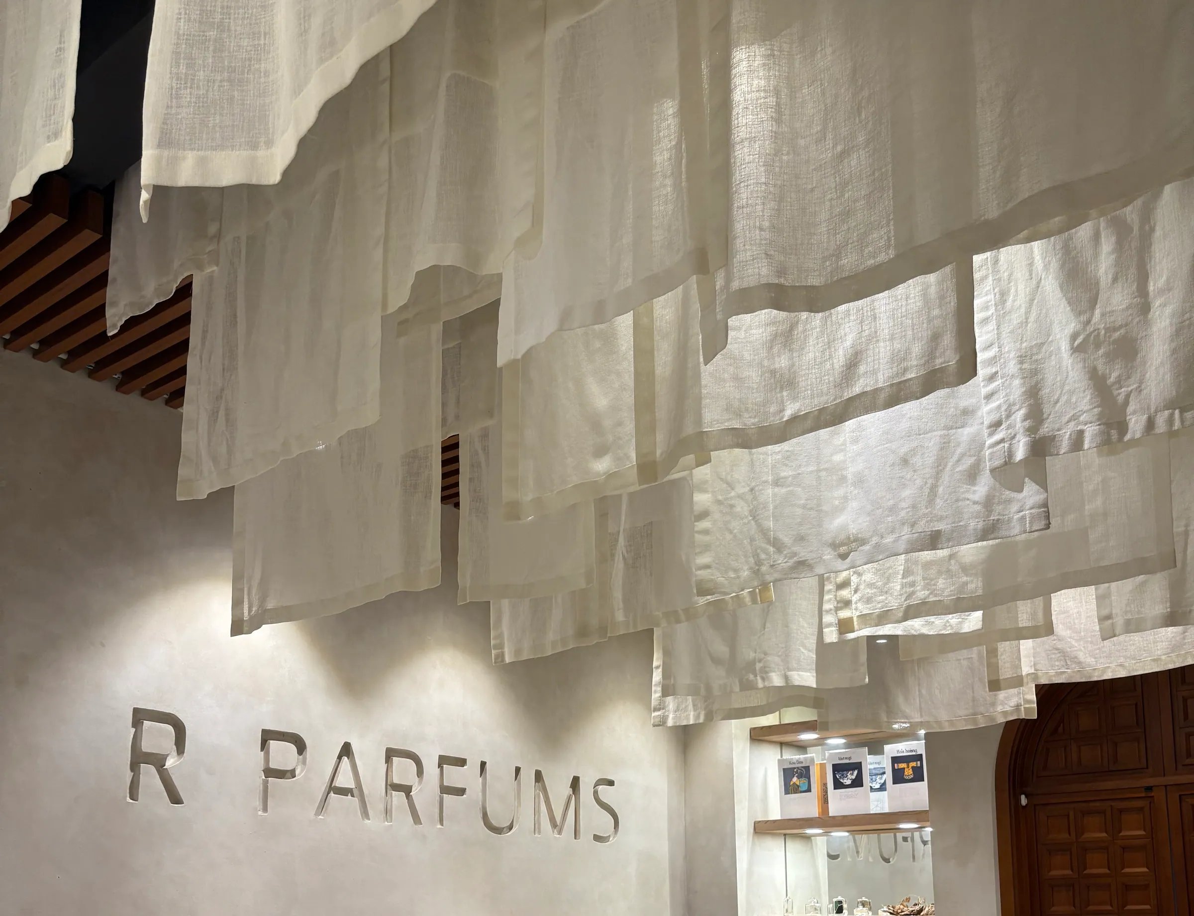 Panel linen tergantung berlapis-lapis dari siling di atas dinding plaster pucat yang diukir huruf R PARFUMS; di bawahnya, sebuah meja konsol kayu yang panjang