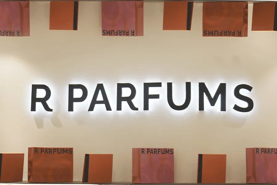 Papan tanda R PARFUMS bercahaya dari belakang pada dinding putih, dibingkai di atas dan di bawah oleh kotak hadiah jingga dan merah jambu