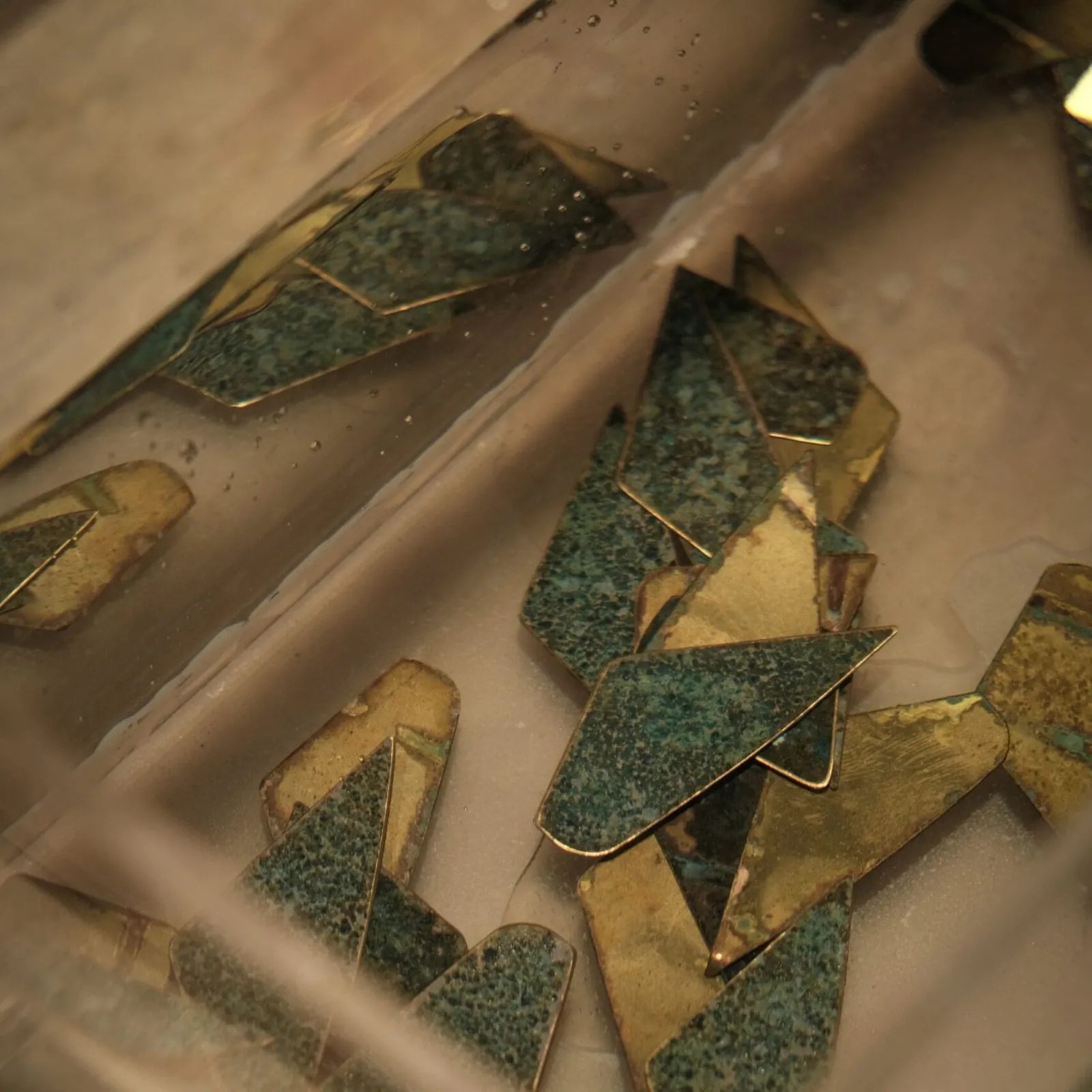 Mga heometrikong piraso ng laton na nakababad sa solusyon, may batik-batik na luntiang patina at ginintuang laton