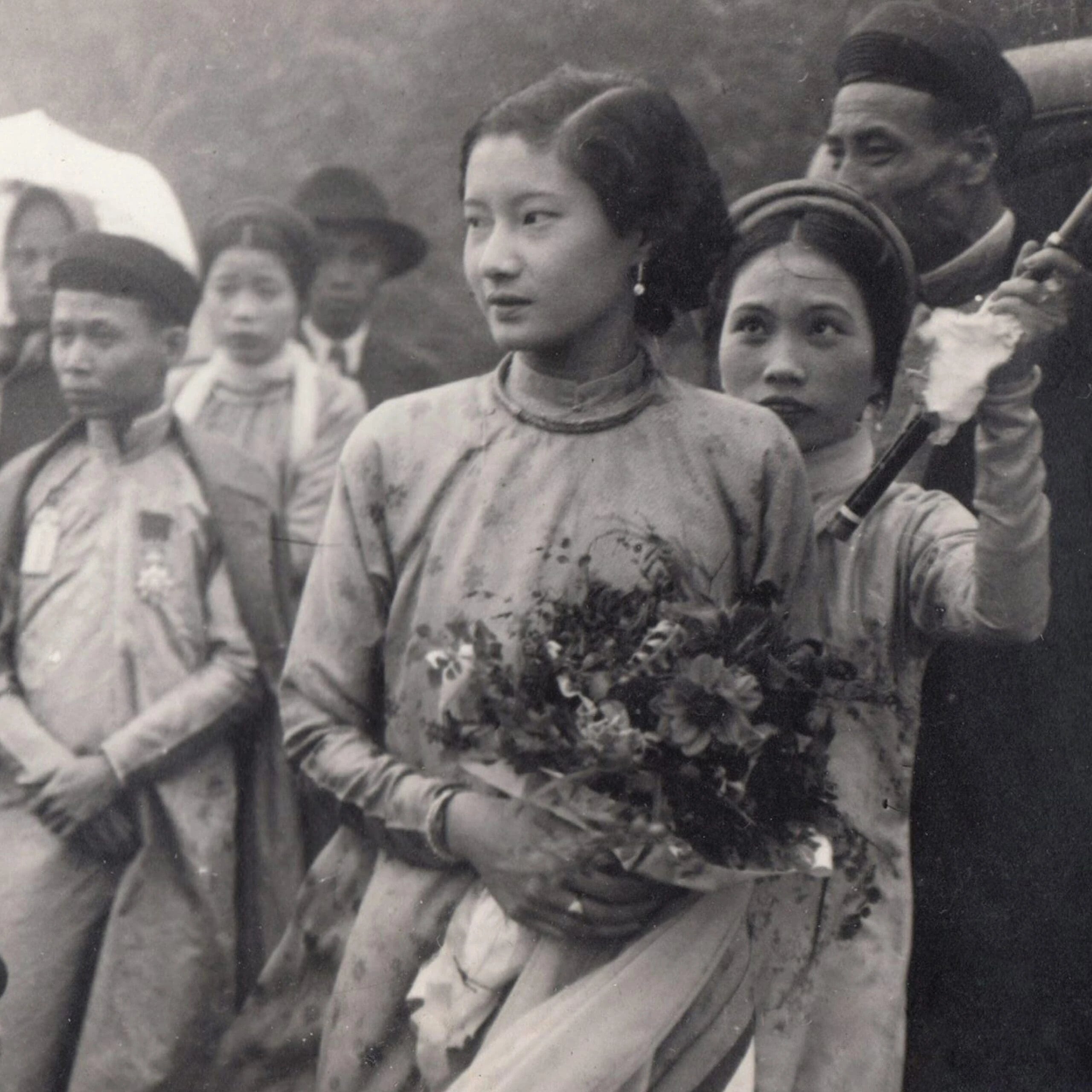 Foto hitam putih lama: seorang wanita muda berbaju áo dài bercorak memegang sejambak bunga, di tengah orang ramai berpakaian tradisional
