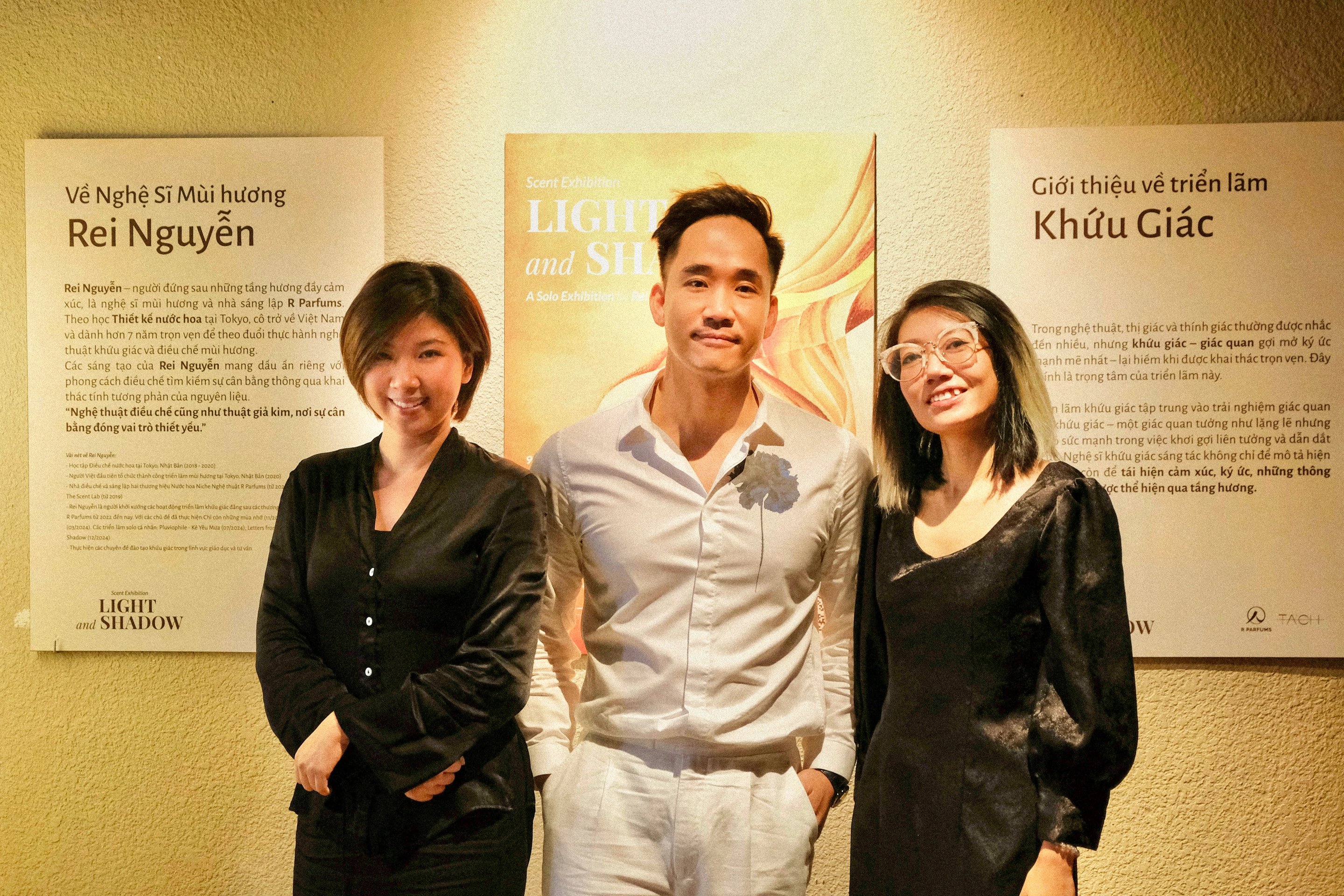 Tiga orang berdiri di hadapan poster Light and Shadow, di antara papan yang memperkenalkan Rei Nguyen dan papan yang memperkenalkan pameran olfaktori