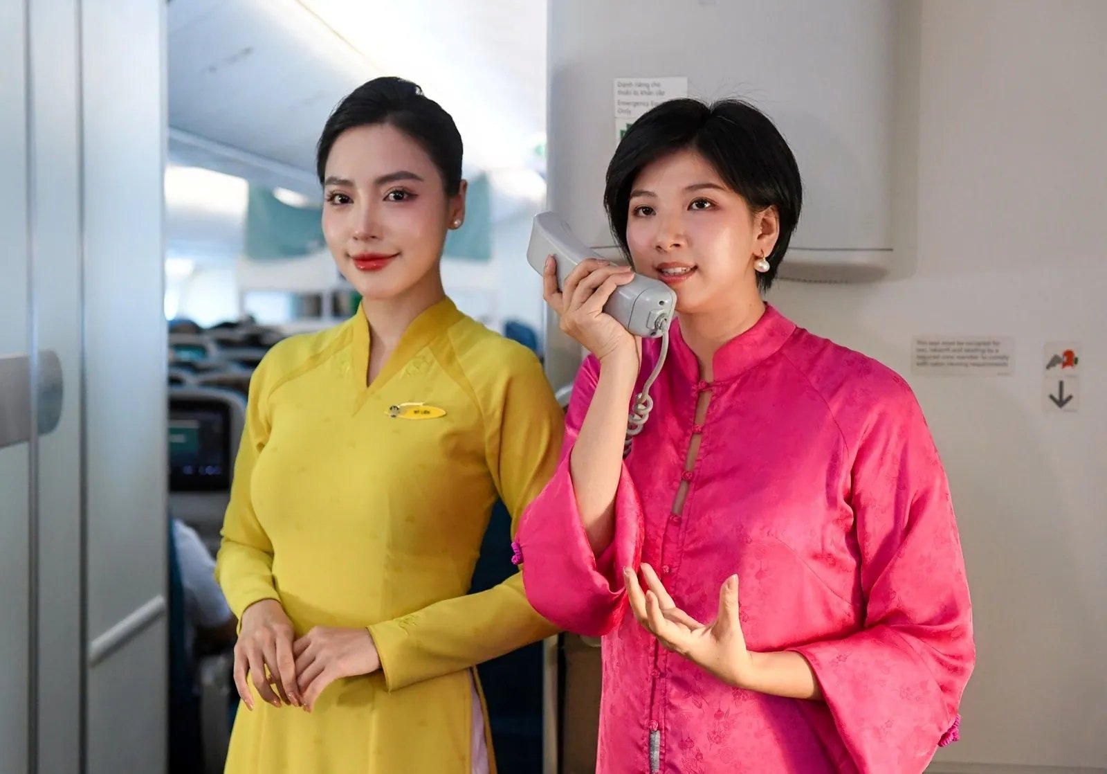 Rei Nguyen ในชุดสีชมพูพูดผ่านอินเตอร์โฟนในห้องโดยสาร ข้างพนักงานต้อนรับบนเครื่องบินในชุด áo dài สีเหลือง