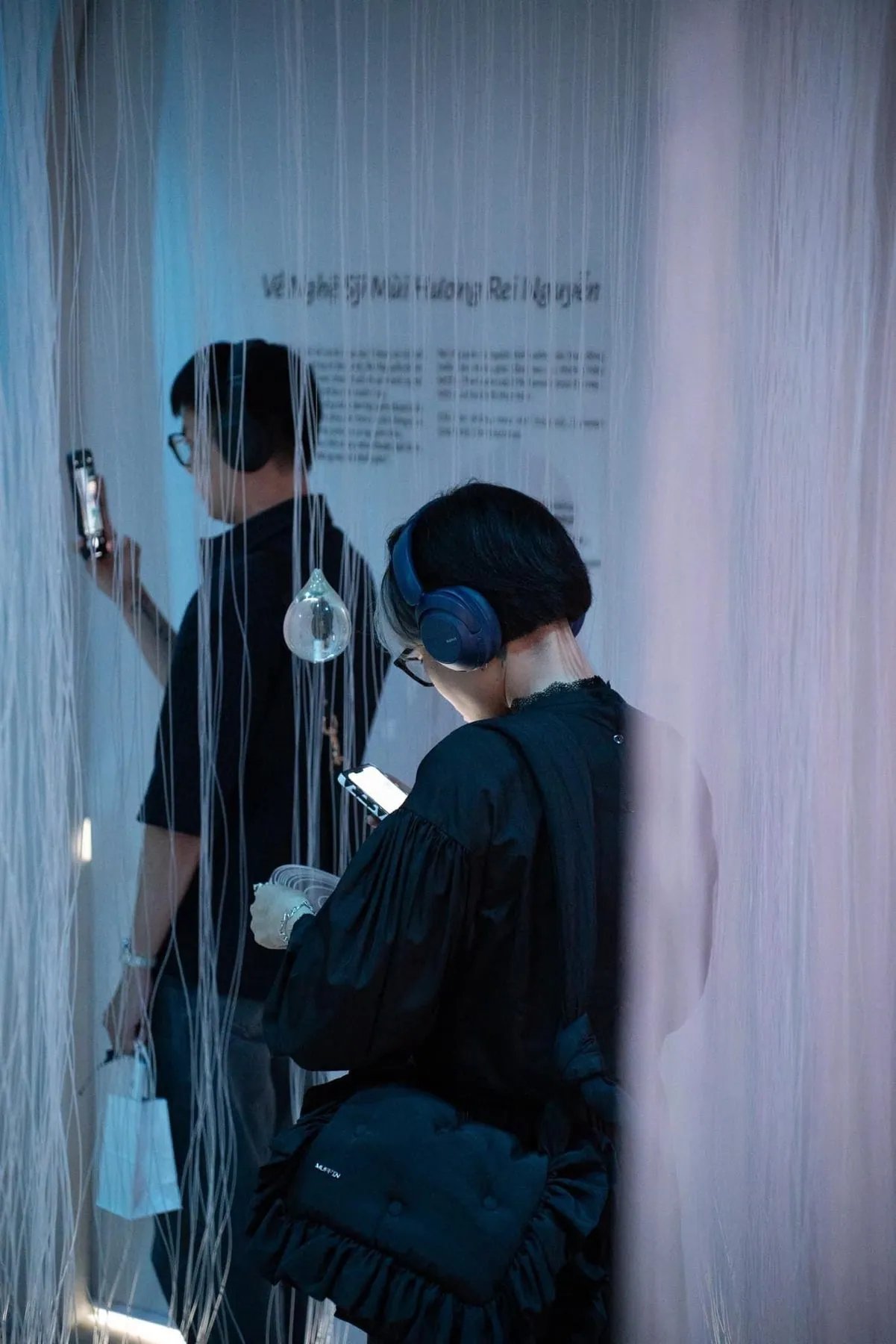 Mga panauhing may headphones na naglalakad sa gubat ng mga puting sinulid at patak ng ulan na salamin, sa eksibisyong Pluviophile