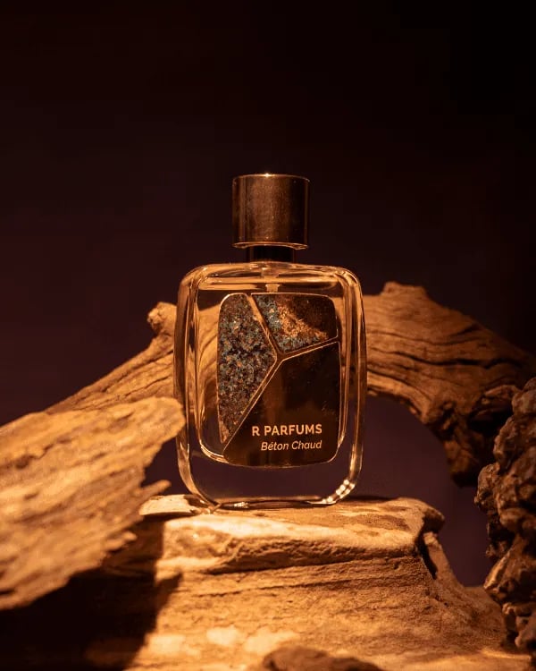 ขวด Béton Chaud ของ R Parfums ที่มีแผ่นทองเหลืองกัดผิวเป็นพาทินาสีเขียวอมฟ้า วางท่ามกลางไม้ลอยน้ำในแสงอบอุ่น
