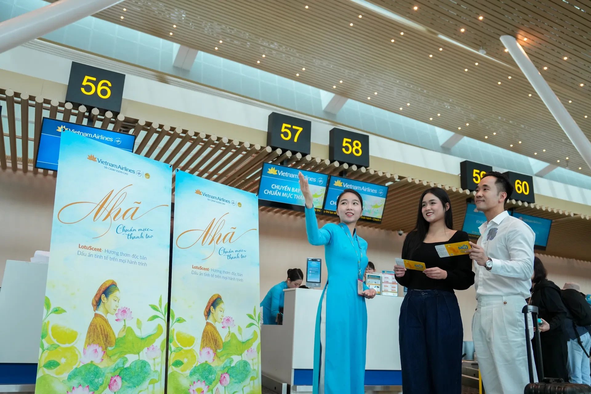 เคาน์เตอร์เช็กอินในสนามบินพร้อมป้ายตั้ง Nhã สองป้าย พนักงานในชุด áo dài สีฟ้ากำลังแนะนำนักเดินทางสองคน