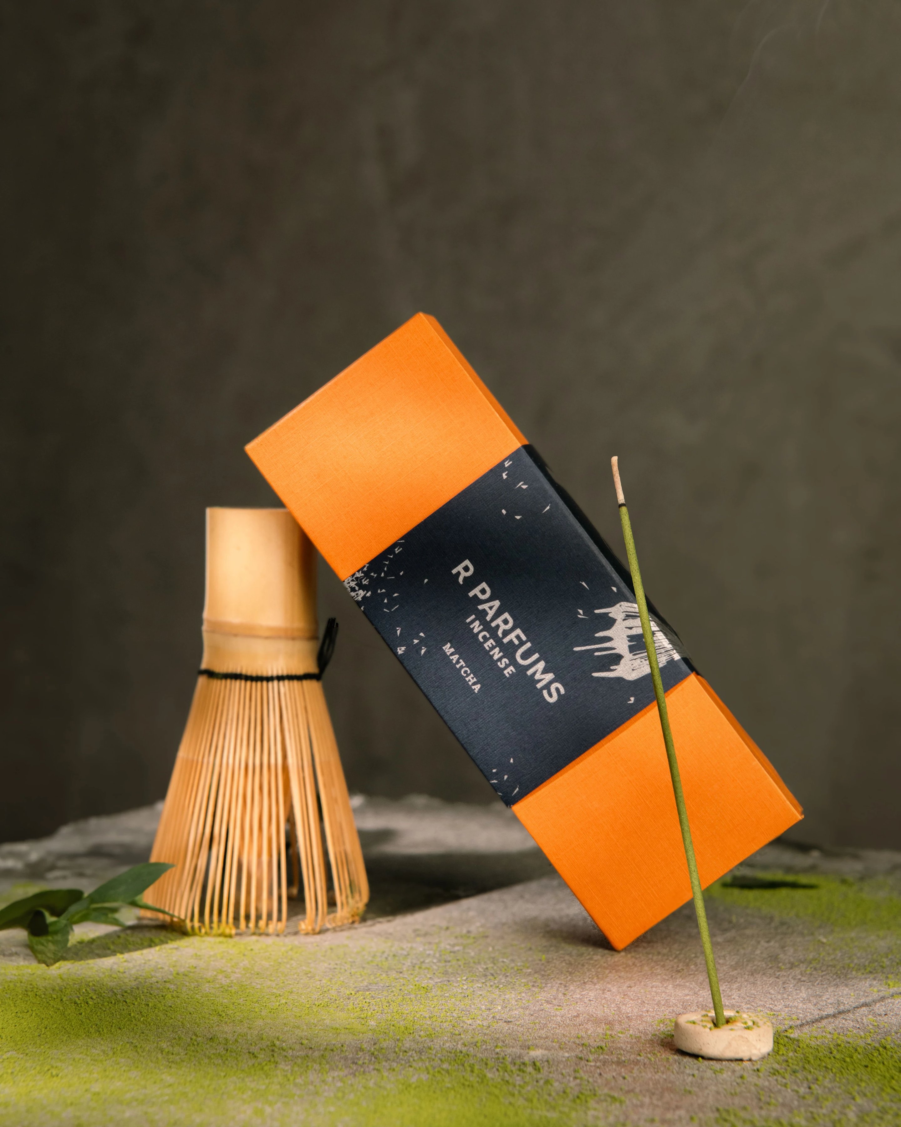Kotak R Parfums Incense Matcha bersandar pada pemukul teh buluh, serbuk matcha bertabur di atas batu
