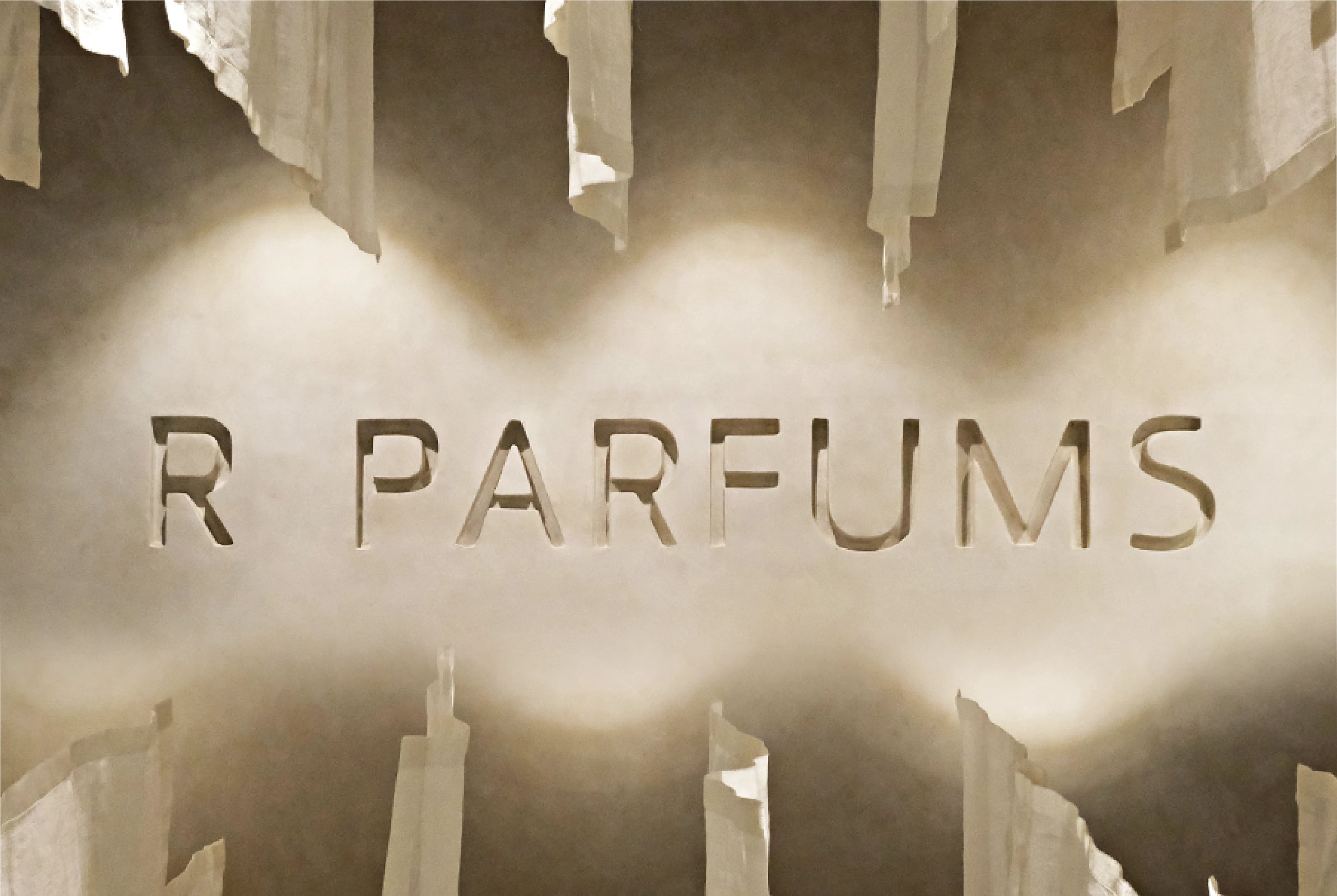 Nama R PARFUMS diukir pada dinding plaster di 34 Nguyễn Duy Hiệu, diterangi dari bawah, dengan jalur linen tergantung di atasnya