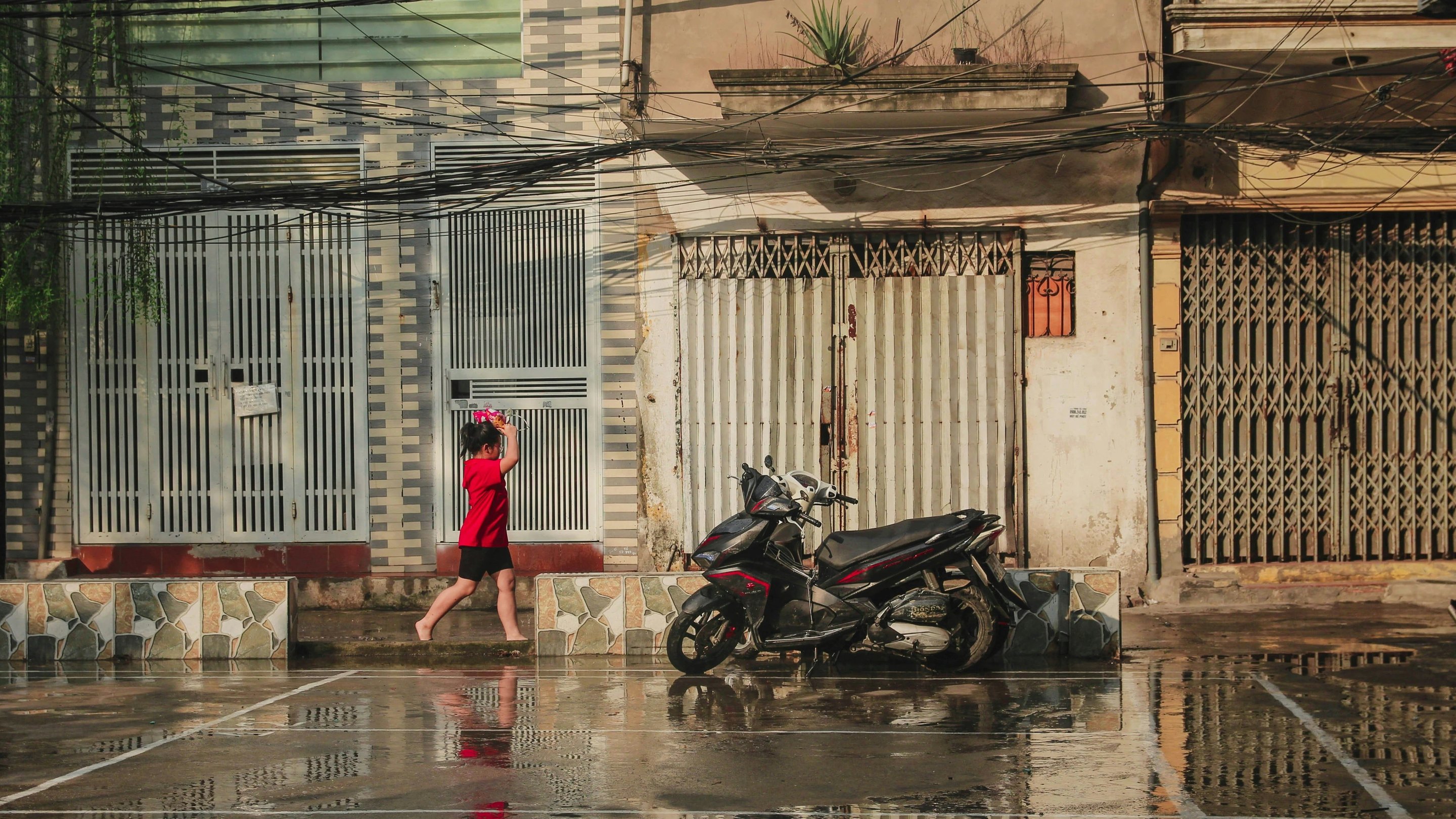Seorang budak perempuan berbaju merah berjalan berkaki ayam di kaki lima yang basah, motosikal diletakkan di hadapan sederet pintu besi gulung di sebuah kejiranan Hanoi selepas hujan
