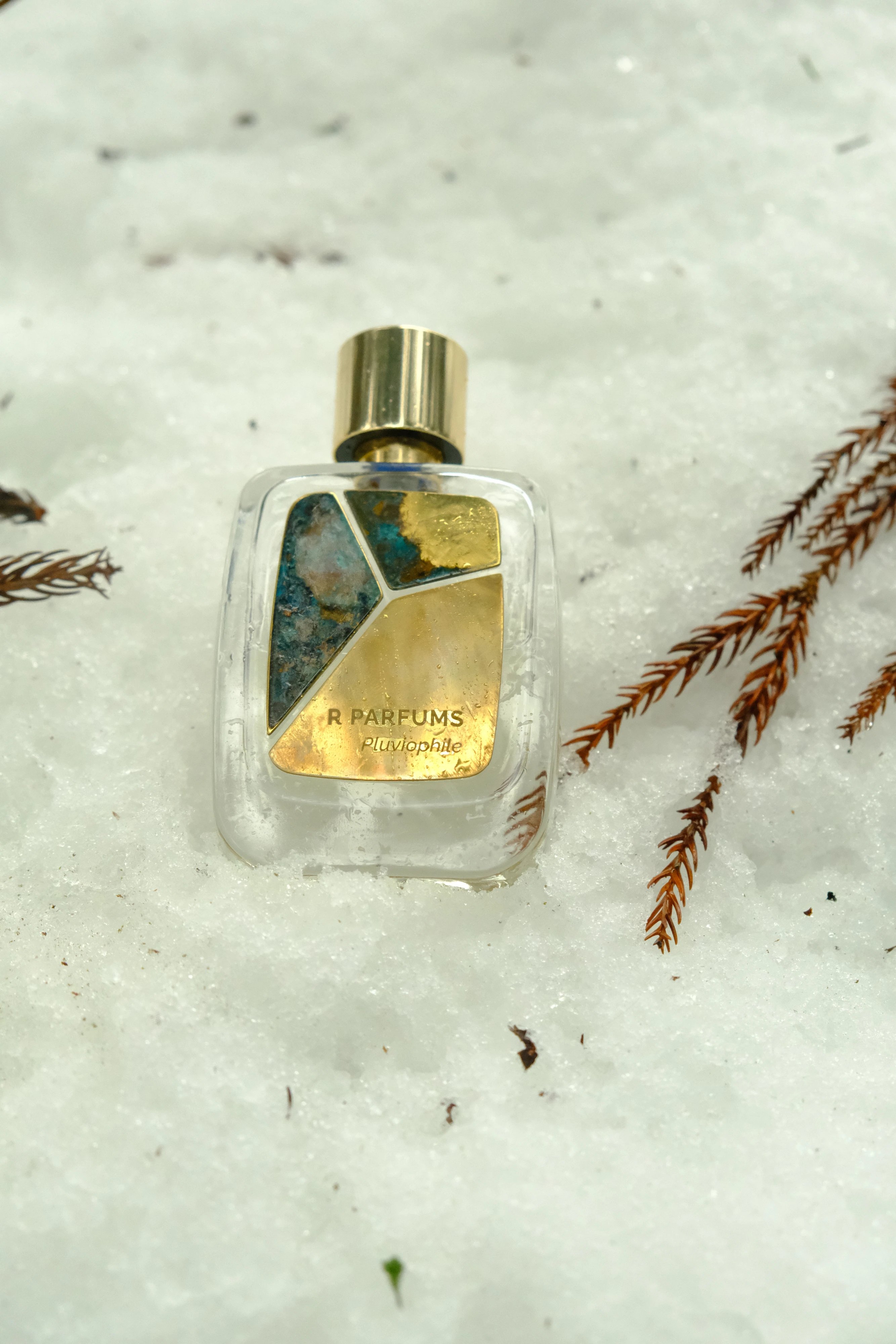 A Pluviophile Patina bottle in snow, close: gold cap, a piece of blue-green patina, a piece half brass and half patina, and a piece of bright brass printed R PARFUMS Pluviophile, sprigs of russet conifer to the right