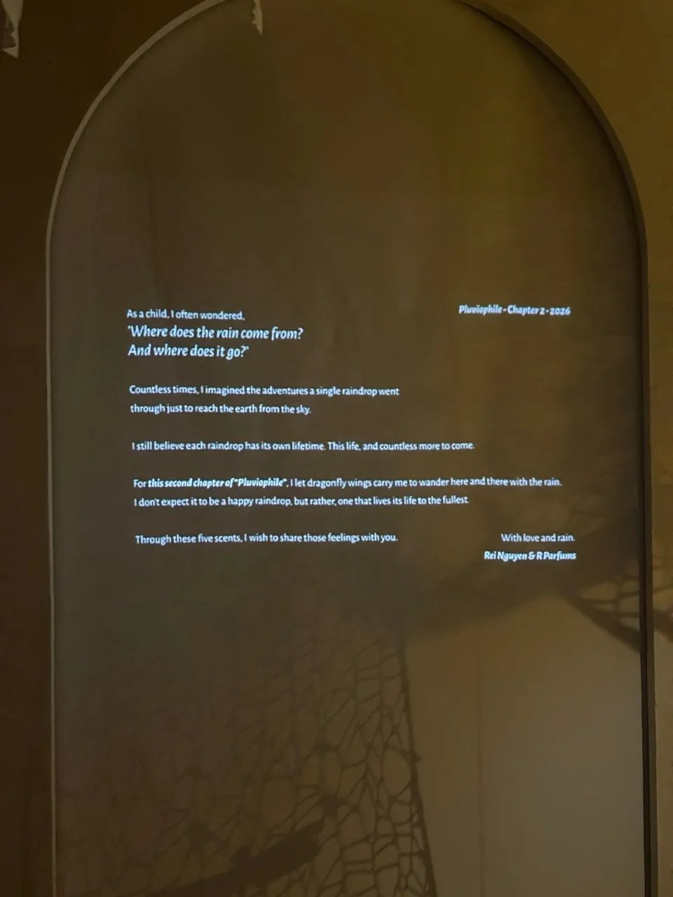 A dark exhibition room: Rei’s letter projected in white on an arched wall, signed Rei Nguyen & R Parfums; the shadow of fine lace falls across the wall
