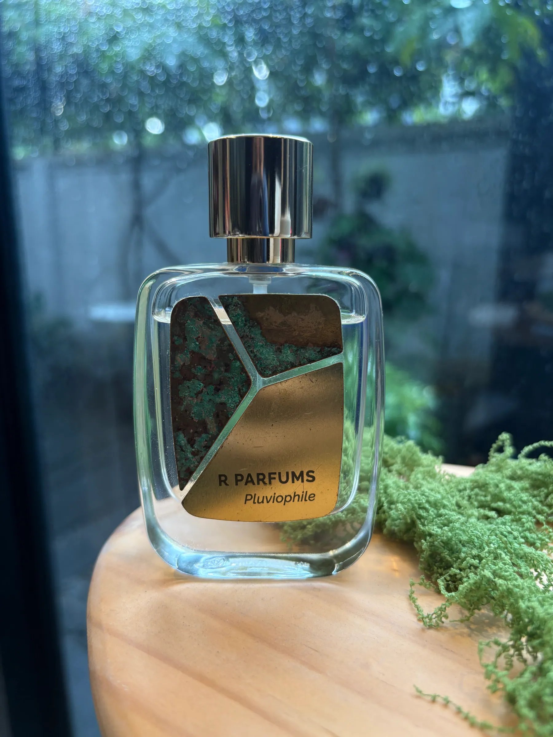 Botol Patina Pluviophile di atas dirian kayu, ‘R PARFUMS Pluviophile’ pada kepingan loyangnya, lumut berjuntai di sisinya, tingkap bergaris hujan di belakang