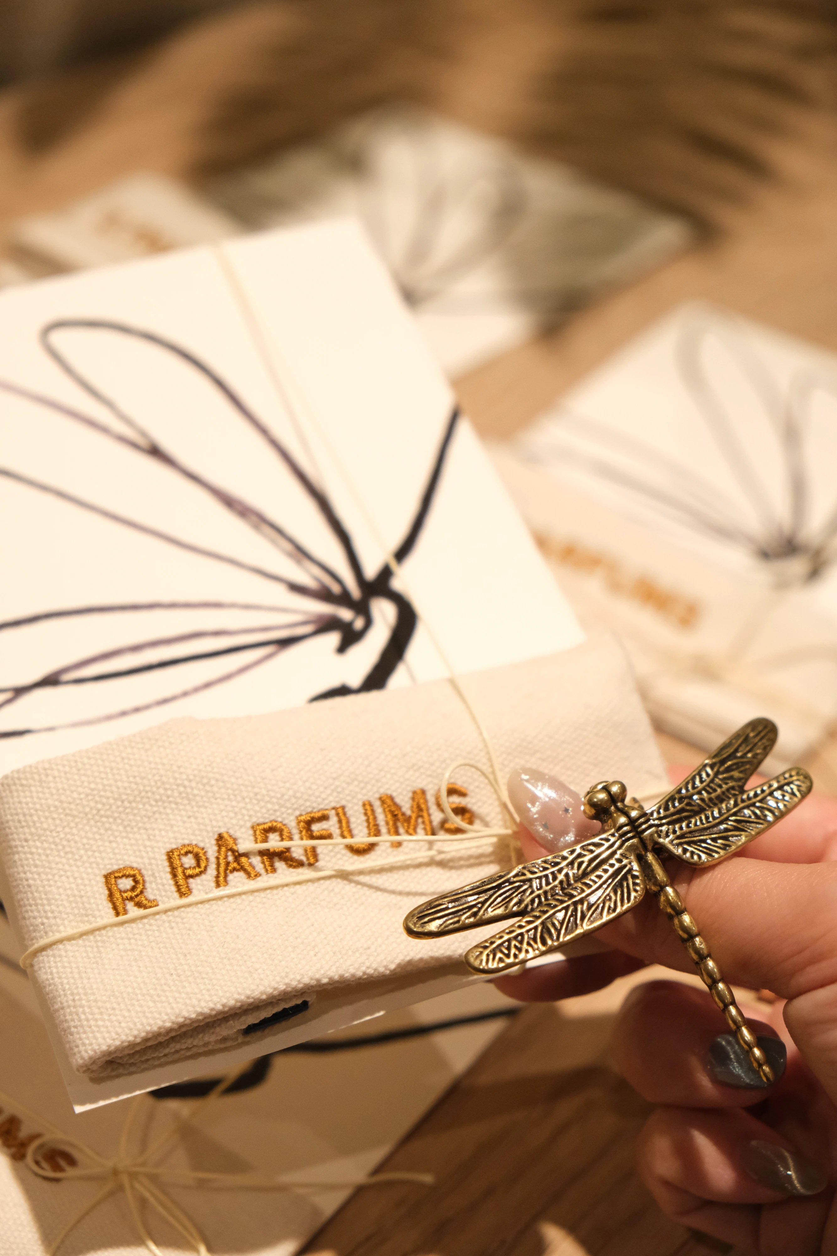 황동 잠자리를 든 손, 그 아래 먹으로 잠자리를 그린 카드와 R PARFUMS 자수 천 주머니