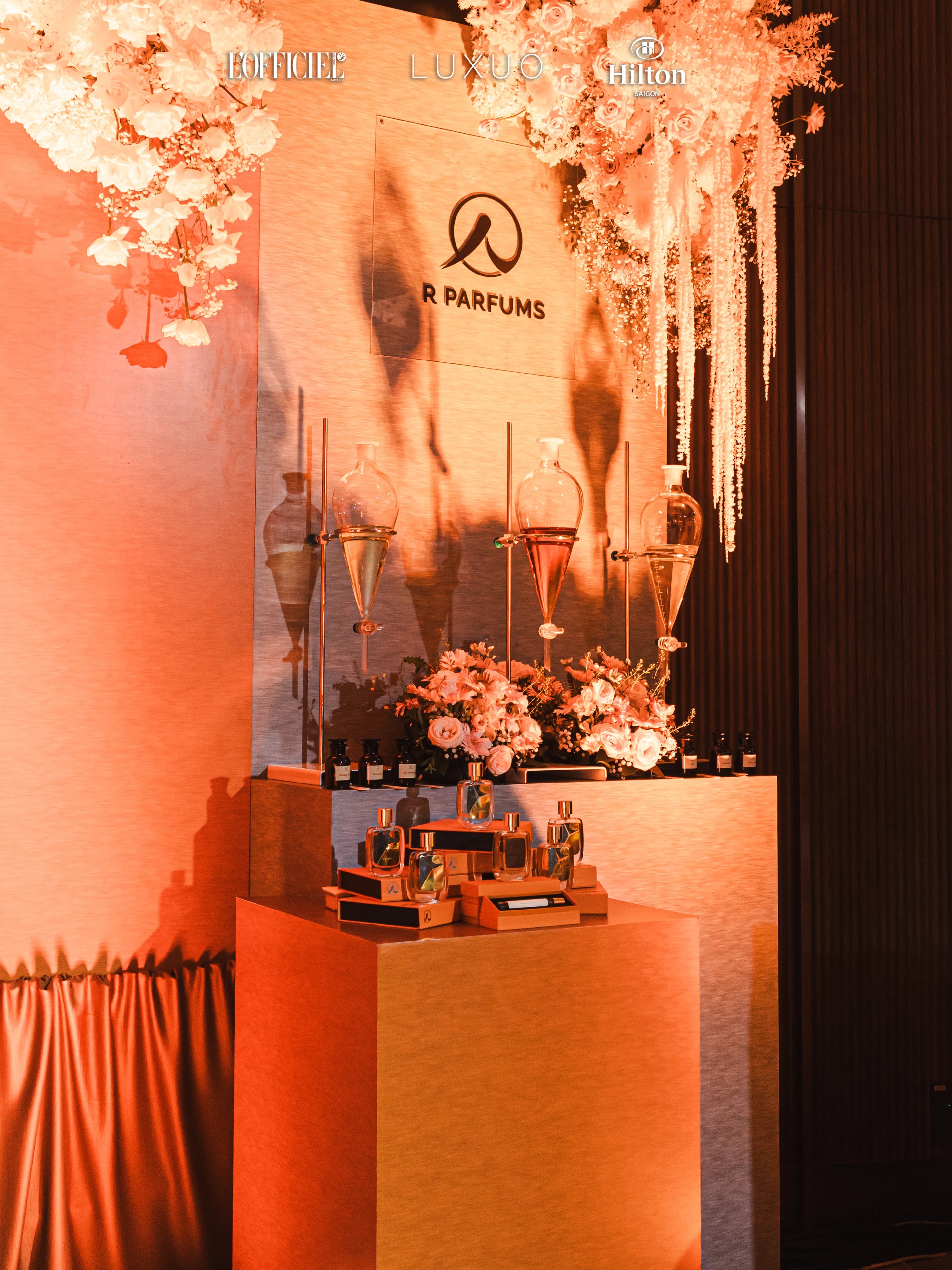 Ang puwesto ng R Parfums sa Wedding Symphony 2025, Hilton Saigon: tatlong embudong salamin sa mga patungang pang-laboratoryo, mga bulaklak na kulay-rosas at puti, mga boteng Patina sa ibabaw ng kanilang kahon